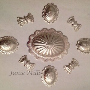 May include: Eight silver-toned metal embellishments with a sunburst design. The embellishments are in various shapes and sizes, including oval, round, and bow-shaped.