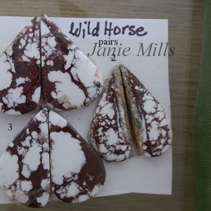May include: Two pairs of Wild Horse cabochons, a type of gemstone, in a heart shape. The cabochons are white with brown and red marbling. The cabochons are labeled with numbers 1 and 3. The text "Wild Horse" and "Jamie Mills" is written on a white card.