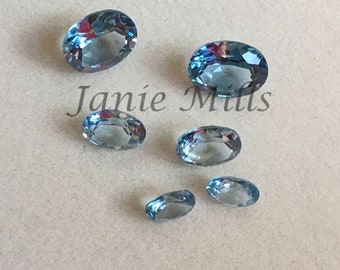 May include: Seven oval-shaped gemstones in various sizes. The gemstones are a light blue color with a slight shimmer. The gemstones are arranged on a white surface.