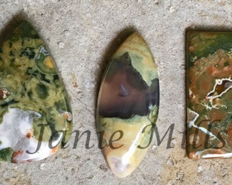 May include: Three polished green, brown, and white jasper cabochons. The cabochons are in the shapes of a teardrop, oval, and rectangle.