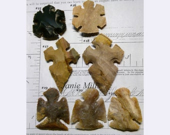 Puede incluir: Una colección de ocho puntas de flecha, algunas con diseños intrincados, mostradas en una superficie blanca con una regla y texto escrito a mano. Las puntas de flecha están hechas de varios tonos de piedra marrón, tostado y negro.