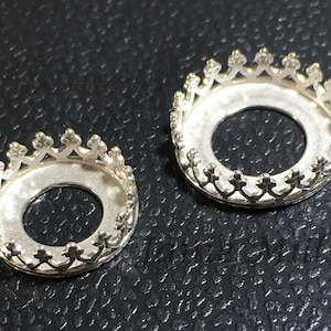 Puede incluir: Dos ajustes de metal de color plateado en forma de corona con una abertura redonda en el centro. Los ajustes están sobre una superficie negra.