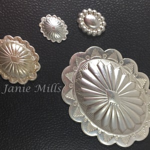 May include: Four silver-toned metal conchos with intricate designs. The largest concho has a scalloped edge and a sunburst pattern. The other three conchos have a more simple design.