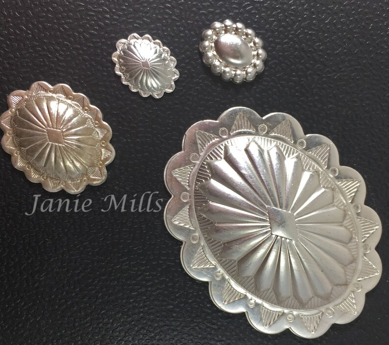 May include: Four silver-toned metal conchos with intricate designs. The largest concho has a scalloped edge and a sunburst pattern. The other three conchos have a more simple design.