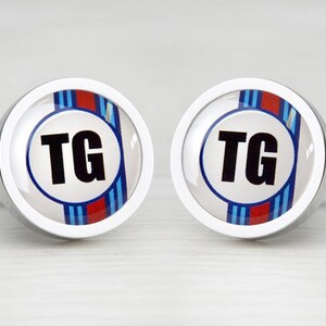 May include: A pair of silver cufflinks with a white background and blue and red stripes. The initials "TG" are in black letters on a white circle in the center of the cufflinks.