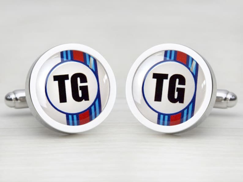 May include: A pair of silver cufflinks with a white background and blue and red stripes. The initials "TG" are in black letters on a white circle in the center of the cufflinks.