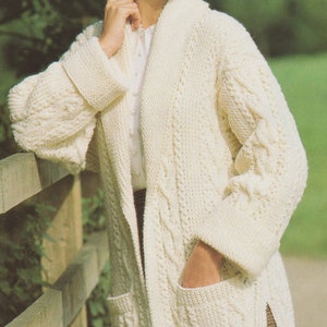 Może przedstawiać: Biały kardigan dzianinowy z kołnierzem szalowym, długimi rękawami i dwiema kieszeniami. Kardigan jest wykonany z dzianiny warkoczowej.