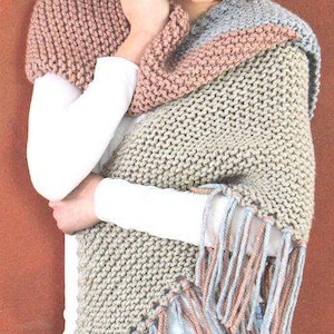 May include: A woman wearing a knitted shawl with a fringed edge. The shawl features a colour block design with shades of pink, grey, and beige. The woman is wearing a white long-sleeved shirt and blue jeans.