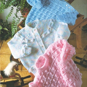 Può includere: Tre maglioni di lana per neonati in diversi colori e motivi. I maglioni sono blu, rosa e bianco con un motivo blu chiaro e bianco. I maglioni sono disposti su una superficie di legno.