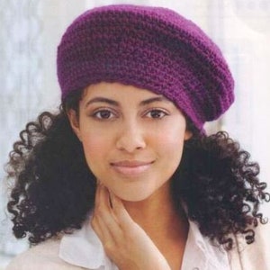 May include: A woman with curly brown hair is wearing a purple knitted beret. The beret is angled on her head, with the brim covering one eye.