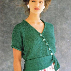 Może przedstawiać: Zielony sweterek z dekoltem w kształcie litery V i krótkimi rękawami. Sweterek zapinany jest na skośne guziki i ma wiązanie w talii.