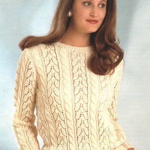 Può includere: Maglione lavorato a maglia color crema con scollo tondo e maniche lunghe. Il maglione presenta un dettagliato motivo a trecce e un motivo a foglie traforate. La modella indossa il maglione con pantaloni grigi.