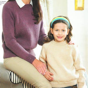 Könnte beinhalten: Eine Frau und ein Kind tragen Strickpullover. Der Pullover der Frau ist tiefviolett, der des Kindes hellbeige. Das Kind trägt ein buntes Stirnband. Die Frau trägt ein weißes Hemd mit Kragen unter ihrem Pullover und eine beige Hose.
