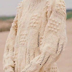 May include: A white knitted sweater with a textured pattern and bobbles. The sweater has a round neckline and long sleeves.