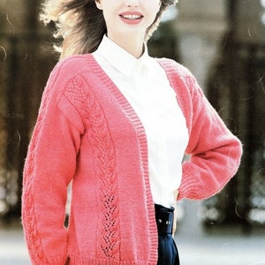 Puede incluir: Un cárdigan de punto rosa con un patrón de trenza y cuello en V. El cárdigan se muestra siendo usado por una modelo con una camisa blanca y pantalones negros. "DOUBLE KNIT" y "28" TO 42" están impresos en la imagen.