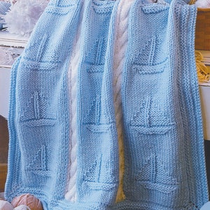 Puede incluir: Una manta de bebé de punto azul claro con un borde de punto de cable blanco. La manta presenta un patrón repetitivo de veleros.