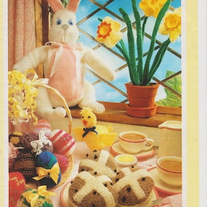 May include: A knitted bunny rabbit, a knitted chick, and a bouquet of knitted daffodils are displayed on a windowsill.  A plate of knitted hot cross buns sits on a table with a cup of tea and a teapot.