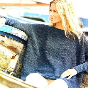 Puede incluir: Una mujer lleva un suéter de punto azul con mangas largas y un corte holgado. Está sentada en un bote de madera con un borde pintado de azul.