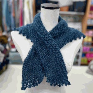 May include: A dark teal knitted scarf displayed on a mannequin. The scarf has a textured, open weave and a scalloped edge. The scarf is draped across the mannequin's shoulders, showcasing its length and design. The background is blurred, suggesting a craft store setting.