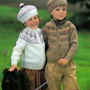 May include: Two children wearing knitted cardigans and hats with bobbles. The girl is wearing a white cardigan with a purple and white pattern and a white hat with a purple bobble. The boy is wearing a brown cardigan with a brown and white pattern and a brown hat with a brown bobble. They are both smiling and looking at the camera.