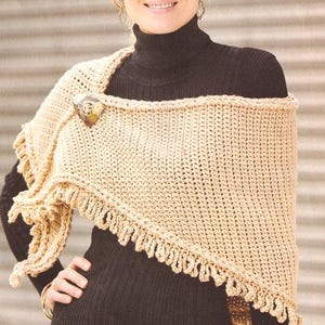 May include: A beige crocheted shawl with a decorative fringe along the edges. The shawl is secured with a decorative brooch. The woman is wearing a black turtleneck sweater.