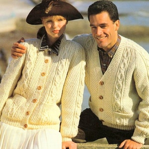May include: A pair of cream-coloured cable knit cardigans with button fastenings. The cardigan on the left has a collar and the cardigan on the right has a V-neck.