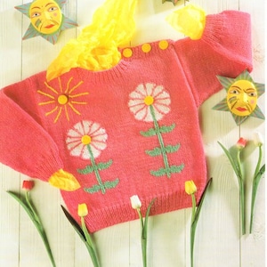 May include: A pink knitted jumper with a floral design featuring white daisies and green stems. The jumper has a round neckline and button fastenings.