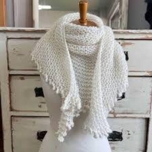 May include: A white, hand-knitted scarf with a textured pattern and a scalloped edge. The scarf is draped over a mannequin head, showcasing its triangular shape and delicate design. The scarf is made of a soft, light-coloured yarn.