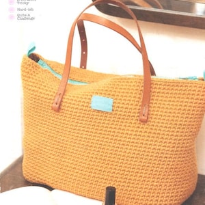 May include: A crocheted tote bag in a warm yellow hue, featuring brown leather handles and a light blue accent patch. The bag has a rectangular shape and a zippered top. The text "Easy Peasy" is visible in the upper left corner.
