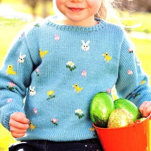 Puede incluir: Un suéter de punto azul claro con cuello redondo y diseños bordados con temática de Pascua, incluyendo conejos, pollitos y flores. El suéter lo lleva puesto un niño que sostiene un cubo rojo lleno de huevos de Pascua de chocolate.