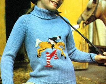 May include: A young person wearing a light blue turtleneck sweater with a knitted design of a horse jumping over a fence. The sweater has a white collar and long sleeves. The person is also wearing a black riding hat and beige pants.