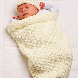 May include: A white crocheted baby blanket with a matching crocheted hat. The baby is swaddled in the blanket and is sleeping peacefully.