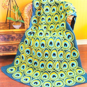 Può includere: Una coperta all'uncinetto fatta a mano a tema pavone. La coperta presenta un design vibrante con colori blu, verde, giallo e nero. Il motivo a piume di pavone è esposto sulla coperta, drappeggiata su una sedia di vimini.