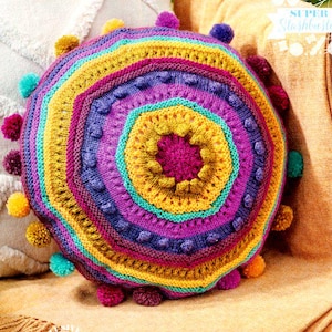 May include: A round, colorful, crocheted pillow with a vibrant, concentric design. The pillow features rings of yellow, purple, teal, and magenta, with textured details and small pom-poms around the edge. The text "SUPER Stashbuster" is visible in the upper right corner.