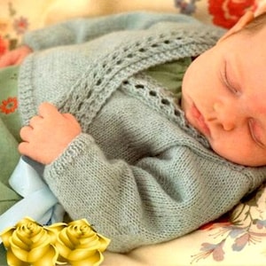 May include: A sleeping baby wearing a light blue knitted cardigan with a delicate lace pattern. The baby is lying on a floral patterned blanket.