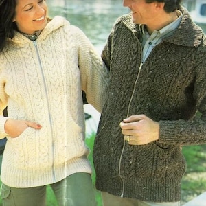 May include: Two people wearing knitted sweaters with zipper closures. The woman's sweater is white and has a hood. The man's sweater is brown and has a collar.