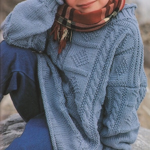 Könnte beinhalten: Ein blauer Strickpullover mit einem Zopfmuster. Der Pullover hat einen runden Halsausschnitt und lange Ärmel. Ein Kind trägt den Pullover und einen rot-schwarzen karierten Schal.