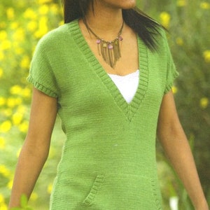 Pode incluir: Um top de malha verde claro com decote em V, mangas curtas e um bolso. O top é usado sobre uma camiseta branca. O decote e a bainha têm um design canelado. A modelo está usando um colar.