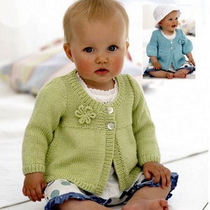 Puede incluir: Un bebé con un cárdigan de punto verde claro con un detalle de flor y dos botones. El bebé también lleva una camisa blanca con ribete de encaje y pantalones cortos de lunares azules y blancos.