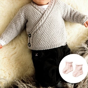 May include: A baby wearing a gray knitted cardigan with a button closure and black pants. The baby is lying on a white fluffy surface. The image also shows a pair of pink knitted baby booties.