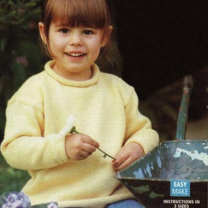 May include: A young girl wearing a yellow knitted jumper and blue jeans holds a white flower. The girl is sitting on a wheelbarrow with a blue and white sign that reads "Easy Make Instructions in 3 Sizes".