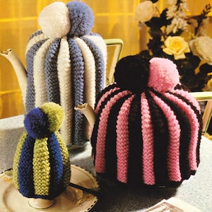 May include: Three knitted teapot and egg cosies. The first is blue and white striped with a white pom-pom on top. The second is black and pink striped with a pink pom-pom on top. The third is green and blue striped with a blue pom-pom on top.