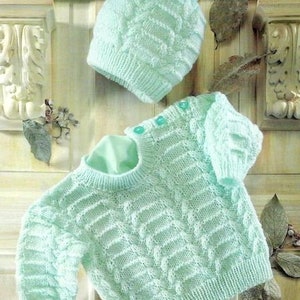 Peut inclure: Un ensemble de tricot pour bébé en vert clair avec un motif en câble. Le pull a un col rond et des manches longues. Le chapeau a un pompon sur le dessus.