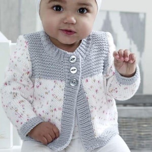 May include: A baby's knitted cardigan in white and gray. The cardigan features a floral pattern on the front and sleeves, with a gray yoke and trim. It has three round buttons down the front. The baby is also wearing a white hat.