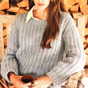 Puede incluir: Un suéter de punto azul claro con textura de canalé. El suéter tiene mangas largas y un cuello redondo. La modelo lleva el suéter con una camisa blanca de cuello debajo.