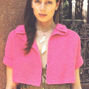 Puede incluir: Una chaqueta de punto rosa brillante, de manga corta y con cuello. La chaqueta tiene cierre de botón y mangas enrolladas. Un collar dorado con colgantes de hojas se lleva sobre una blusa estampada.