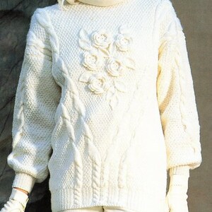 May include: White knitted sweater with a cable knit pattern and a floral design on the front. The sweater has a turtleneck and long sleeves.