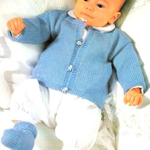 Op de afbeelding: Een baby draagt een lichtblauwe gebreide trui met knopen. De trui heeft een witte kraag en de baby draagt een witte broek.
