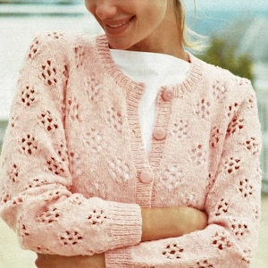 May include: A pink crocheted cardigan with a button-up front and a delicate openwork pattern. The cardigan is worn over a white shirt.