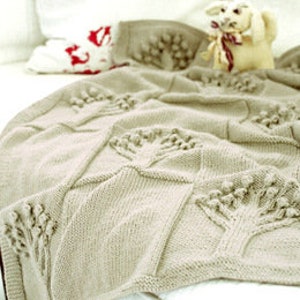 May include: A beige knitted blanket with a tree pattern. The blanket is folded over a bed, with a stuffed animal and pillows in the background. The tree pattern is raised and textured.